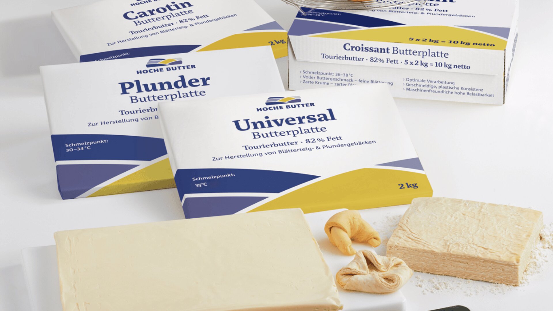 Auf einer weißen Fläche sind Blöcke verpackter Hoche-Butter-Produkte mit den Bezeichnungen Carotin, Plunder und Universal neben unverpackten Butterblättern und Stücken von rohem Gebäckteig ausgestellt.