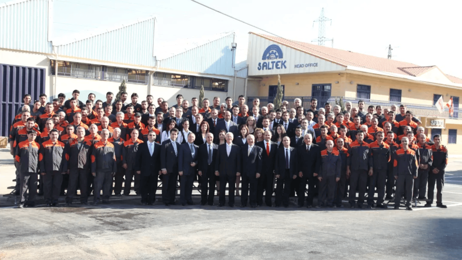 A large group of people, some in black suits and others in black and red uniforms, pose for a group photo outside the SALTEK head office building on a sunny day.