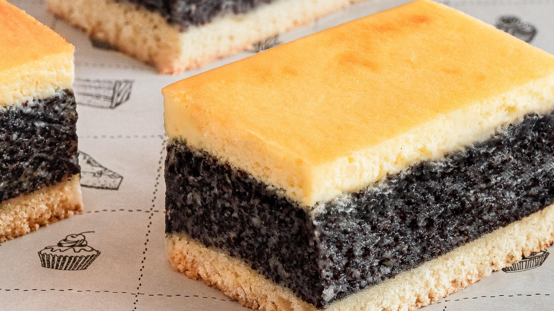 Eine Nahaufnahme von geschichteten Kuchenstücken mit einer hellgelben Oberseite, einer dicken dunklen Mittelschicht und einer hellen unteren Kruste, platziert auf gemustertem Papier mit kleinen Dessertillustrationen.