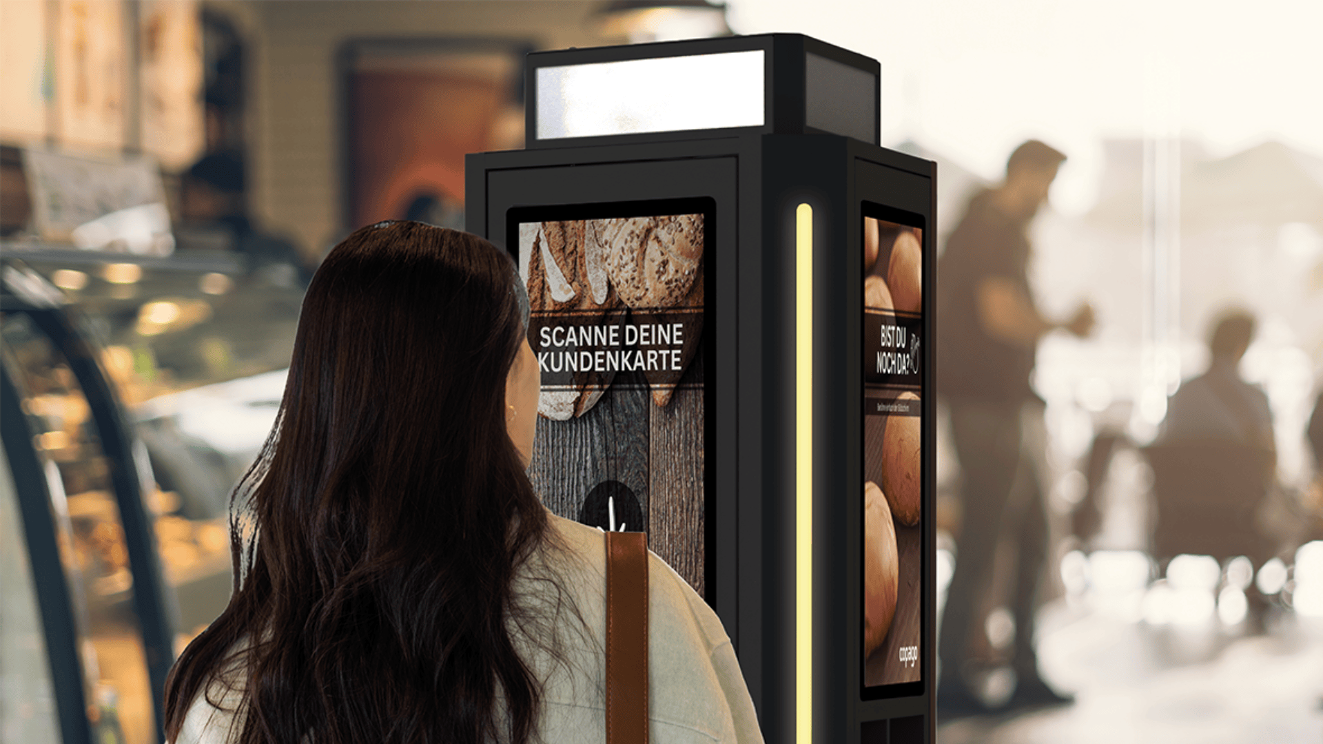 Eine Frau mit langen dunklen Haaren steht vor einem Selbstbedienungskiosk in einer Bäckerei oder einem Café. Auf dem Bildschirm des Kiosks erscheint auf Deutsch die Aufforderung, eine Kundenkarte zu scannen. Im Hintergrund sind weitere Personen unscharf zu sehen.