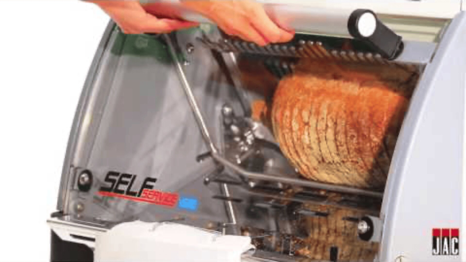 A person lifts the cover of an automatic bread slicer labeled SELF to load a loaf of bread inside the machine. A sliced loaf is partially visible. The JAC logo is seen in the bottom right corner.