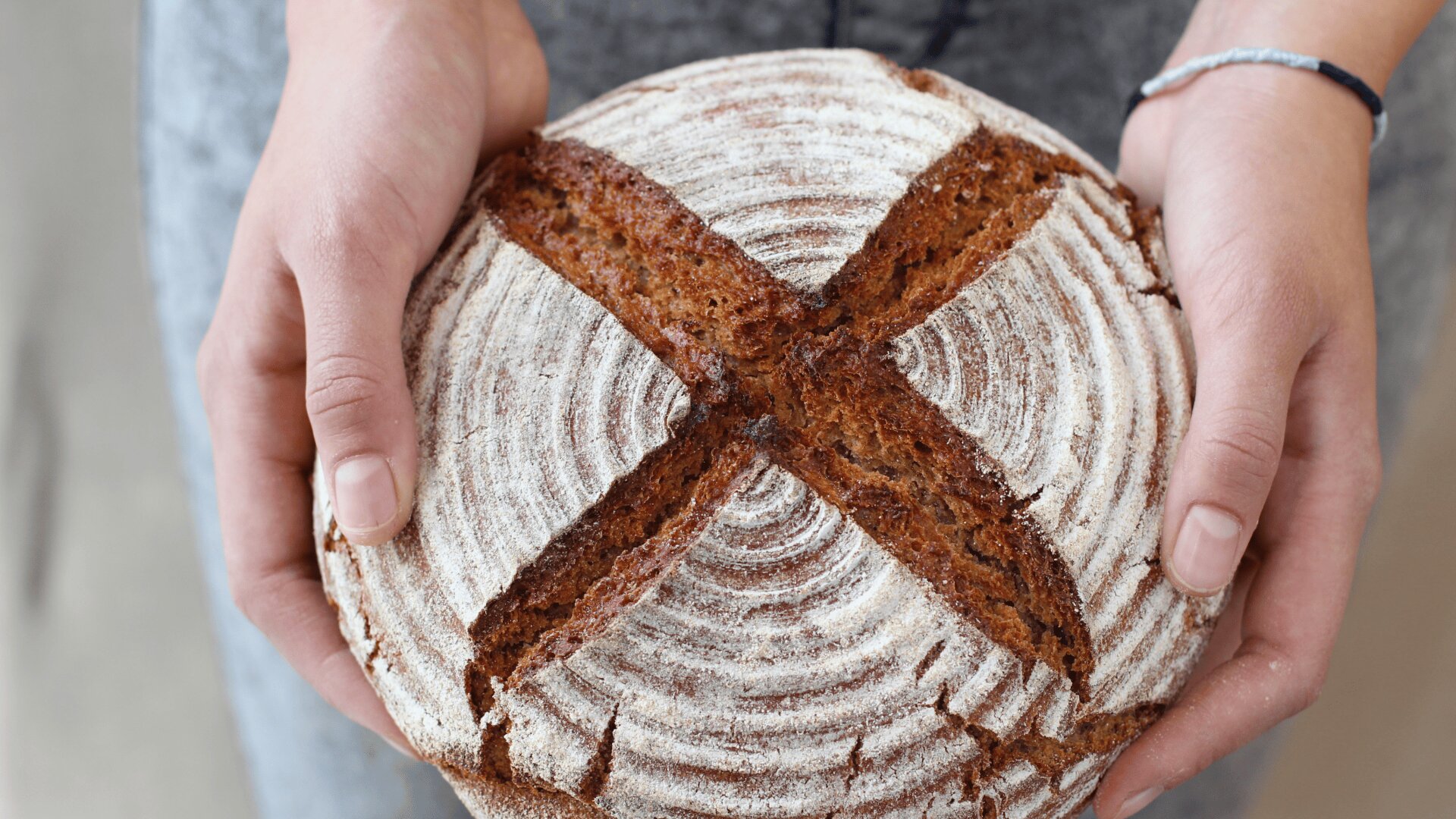 Eine Person hält einen runden Laib handwerklich hergestellten Brotes mit einem spiralförmigen Muster und einem großen X in der Kruste, von oben gesehen.