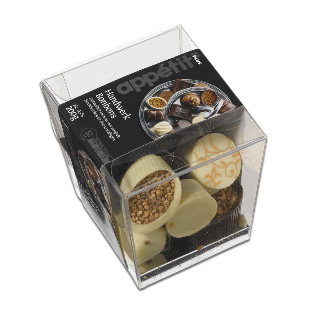 Eine durchsichtige Plastikschachtel mit verschiedenen handgefertigten Bonbons mit einem schwarzen Etikett auf der Oberseite, auf dem appetit Handwerk Bonbons steht. Die Schachtel enthält verschiedene weiße und dunkle Pralinen mit dekorativen Toppings.