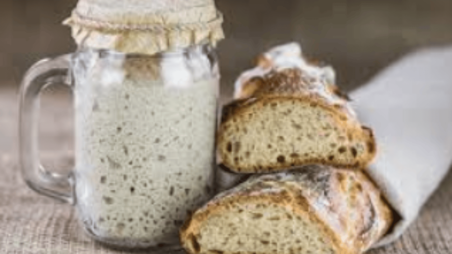 Ein Glas mit blubberndem Sauerteigstarter steht neben zwei in ein Tuch eingewickelten Scheiben rustikalen Sauerteigbrots auf einer Holzplatte.