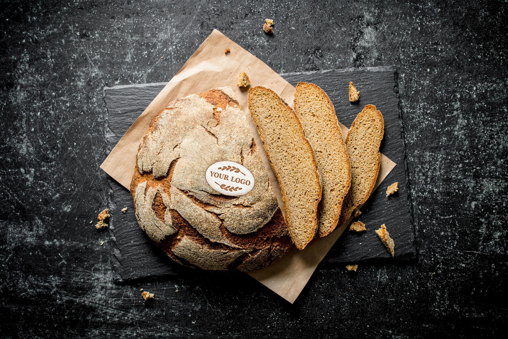 Round rustic loaf of bread on parchment paper with three slices cut, placed on a dark slate surface. A small label reading Your Logo is on top of the loaf. Scattered crumbs surround the bread.