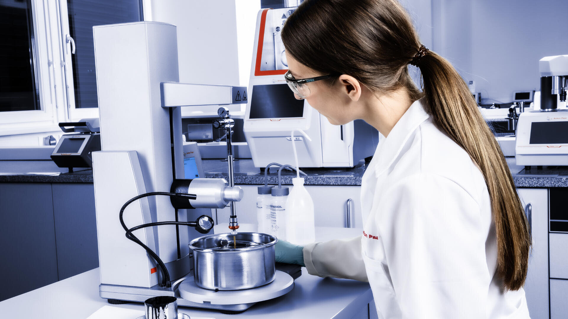 A scientist in a white lab coat and safety glasses uses laboratory equipment to conduct an experiment, mixing substances in a metal container inside a modern laboratory setting.