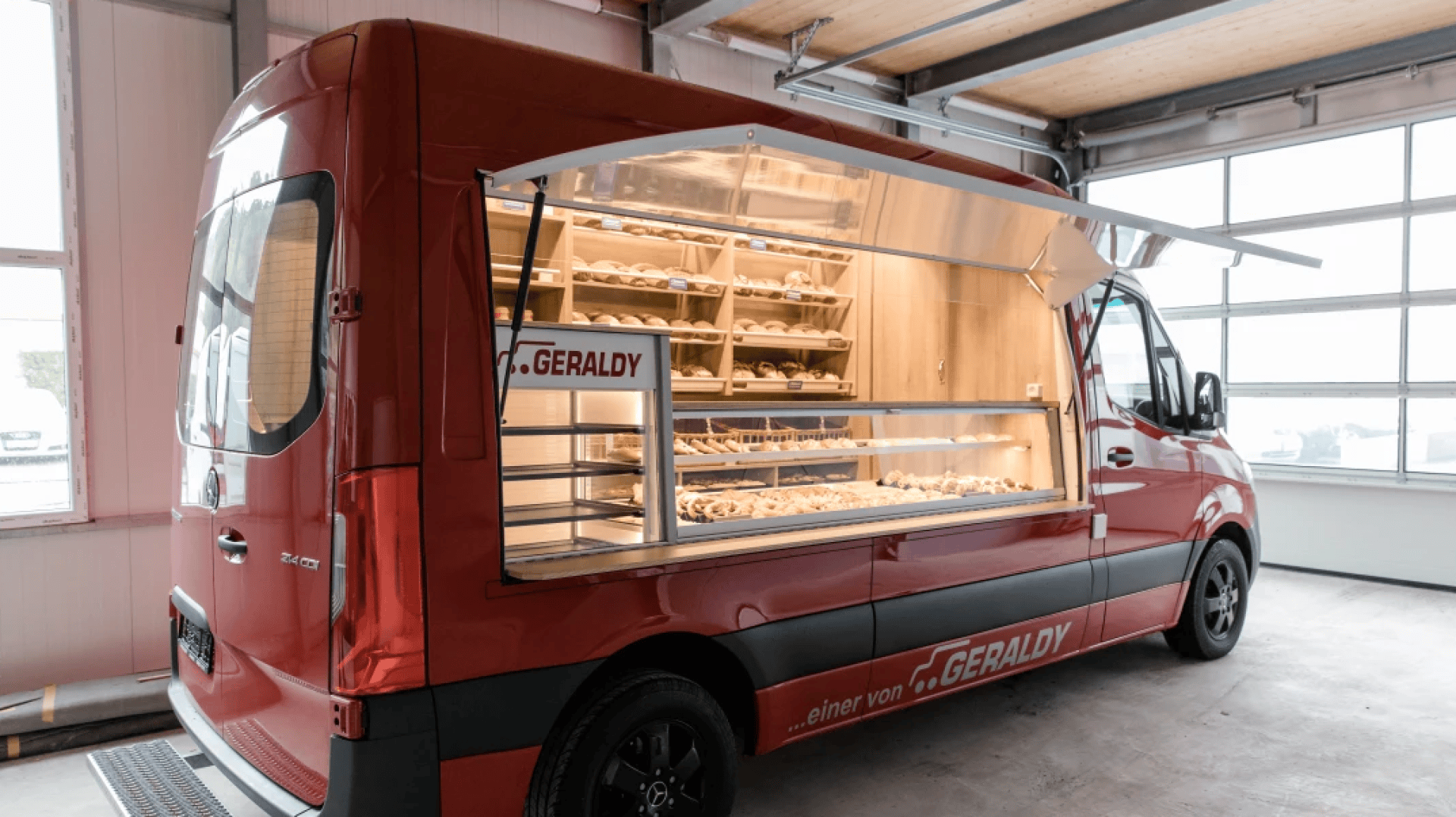 Ein roter Imbisswagen mit einem offenen Seitenfenster bietet Regale mit verschiedenen Brotsorten und Gebäck an. Der Wagen ist in einer Garage mit großen Fenstern geparkt, die natürliches Licht hereinlassen.