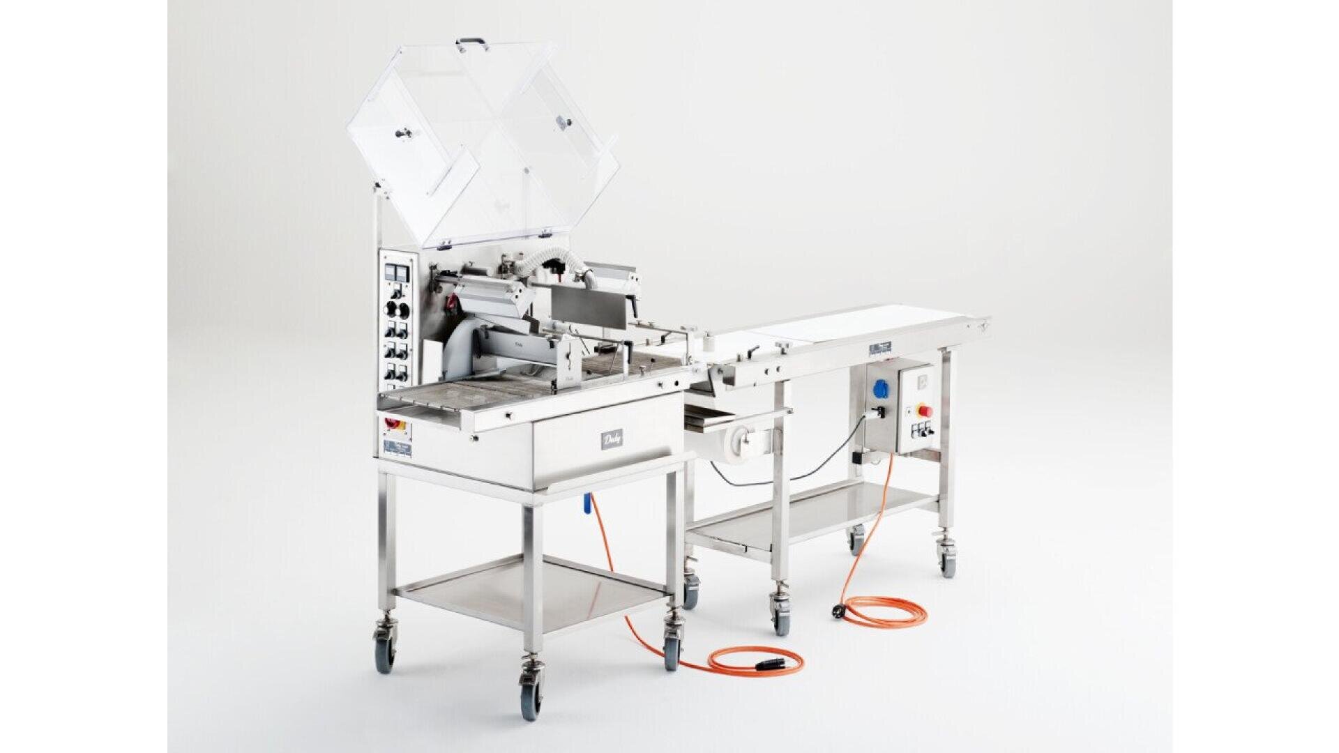 A stainless steel industrial food processing machine with a conveyor belt, control panel, and open lid, standing on wheels with orange power cables on the floor against a plain white background.