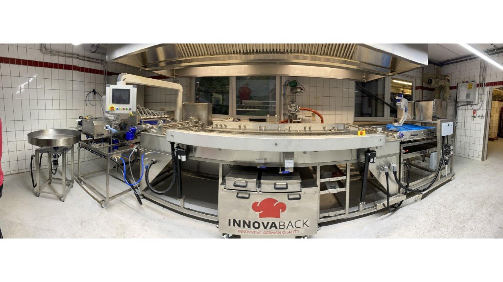 A panoramic view of a modern, automated bakery production line in a clean, tiled room, with stainless steel machinery and a conveyor belt labeled INNOVABACK.