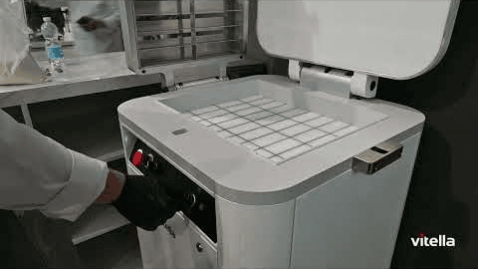 Eine Person, die einen schwarzen Handschuh trägt, bedient in einem Labor oder einer Küche eine weiße Maschine mit einer Gitterfläche und einem offenen Deckel. Das Logo vitella ist in der unteren rechten Ecke zu sehen.