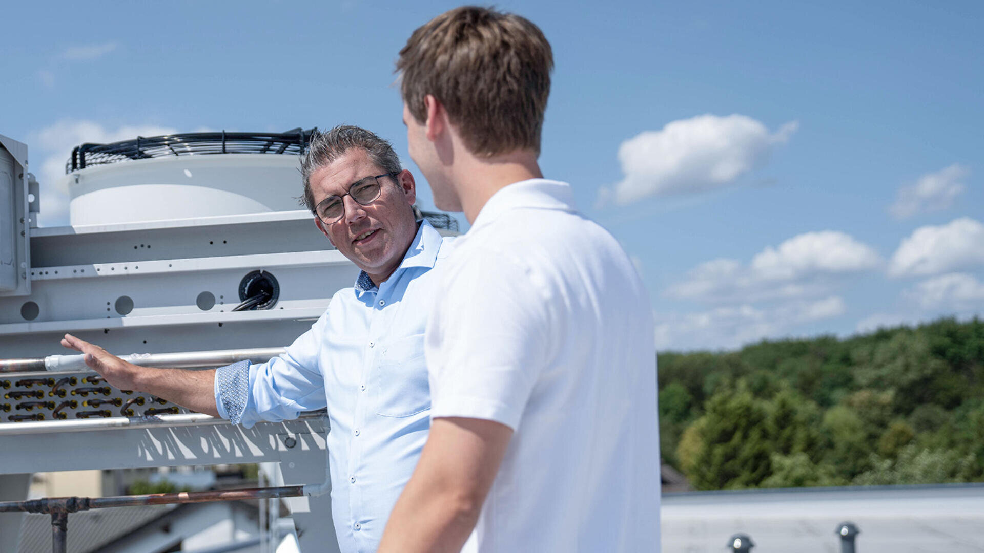 Zwei Männer stehen auf einem Dach neben einer Industrieanlage und unterhalten sich an einem sonnigen Tag mit blauem Himmel und Wolken. Ein Mann gestikuliert in Richtung der Maschinen, während der andere zuhört. Im Hintergrund sind Bäume zu sehen.