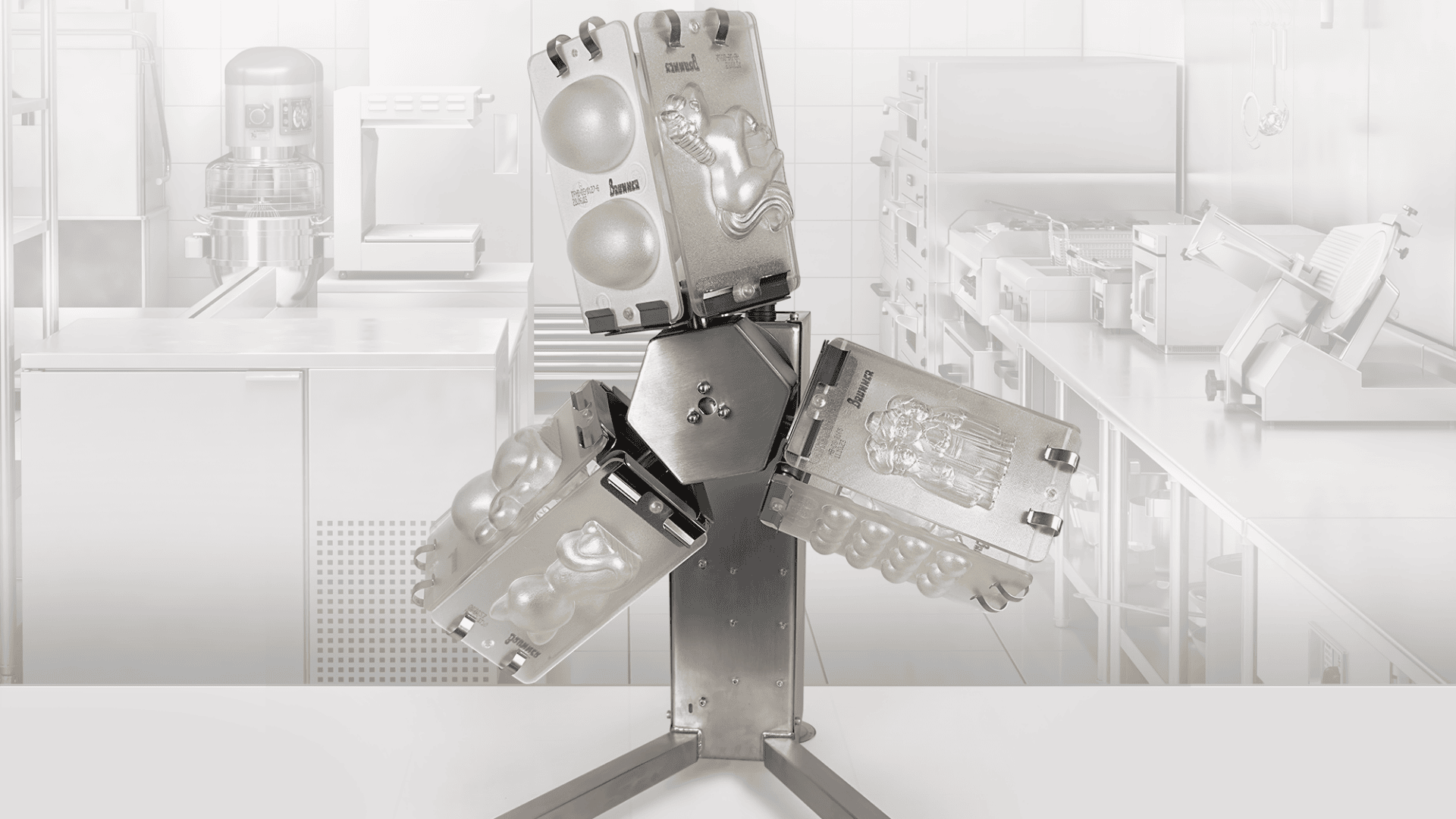 Eine Metallmaschine mit vier aufklappbaren Formen, die jeweils für die Herstellung verschiedener Schokoladenfiguren geeignet sind, steht in einer Großküche mit Edelstahlgeräten im Hintergrund.