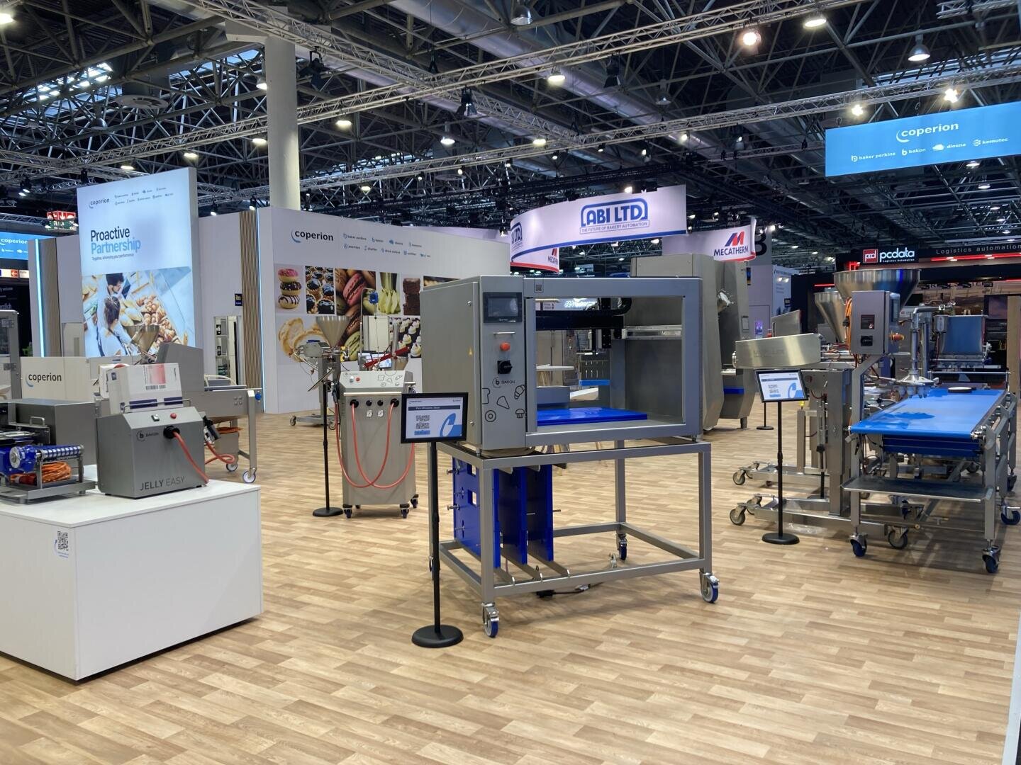 Ausstellungshalle mit verschiedenen ausgestellten industriellen Lebensmittelverarbeitungsmaschinen, darunter Förderbänder und Bedienfelder, umgeben von Firmenständen und Bannern unter heller Beleuchtung.