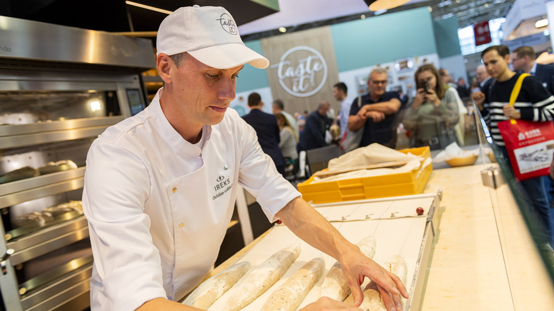 Ein Bäcker in weißer Uniform formt Baguettes auf einer hölzernen Theke bei einer Bäckereiveranstaltung, während im Hintergrund Menschen zusehen und hinter ihm Öfen zu sehen sind.