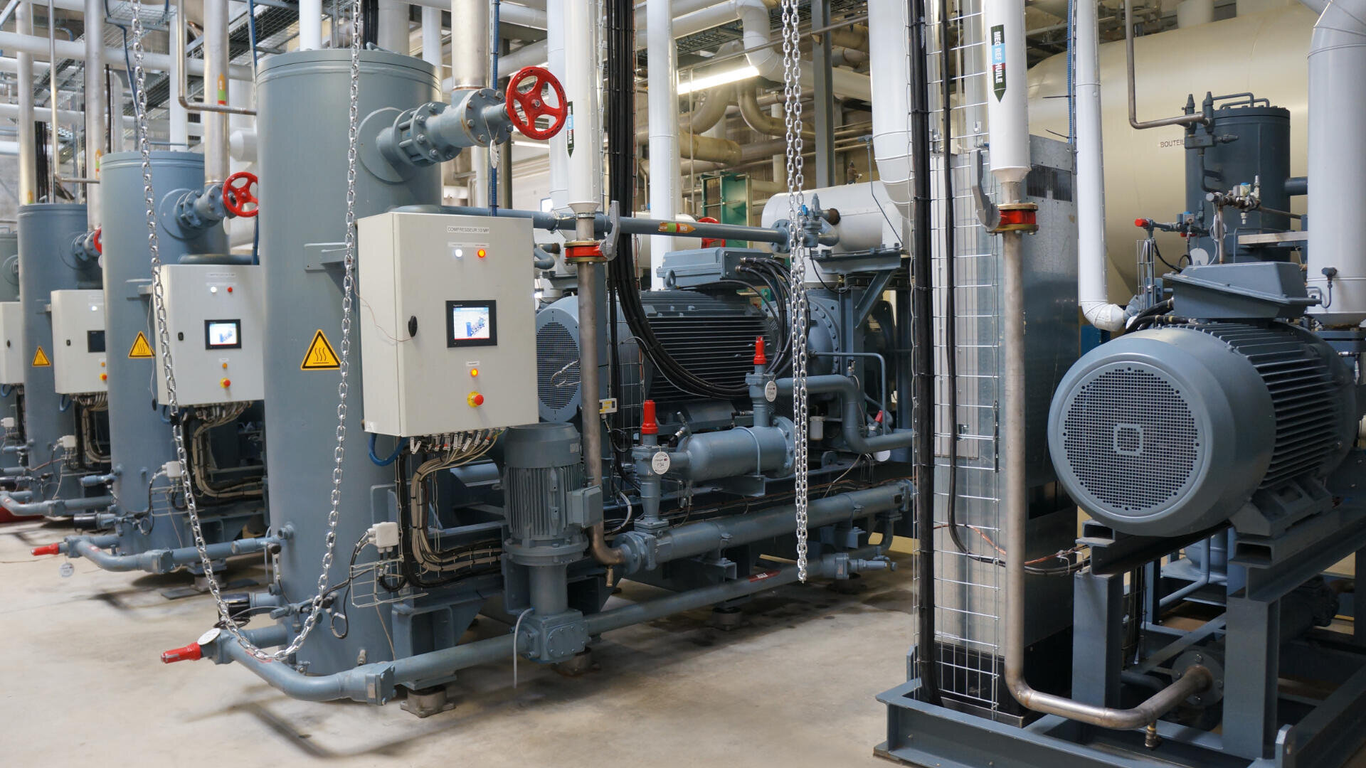 Rows of large industrial machines with control panels, pipes, and chains in a factory setting; electrical warning signs are visible on several units. The equipment is clean and organized.