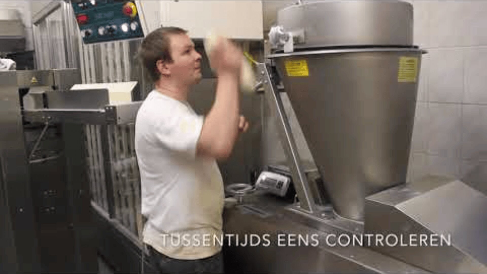 A man in a white shirt operates a large industrial mixing machine in a bakery or food production facility. Dutch text at the bottom reads, “TUSSENTIJDS EENS CONTROLEREN.”.