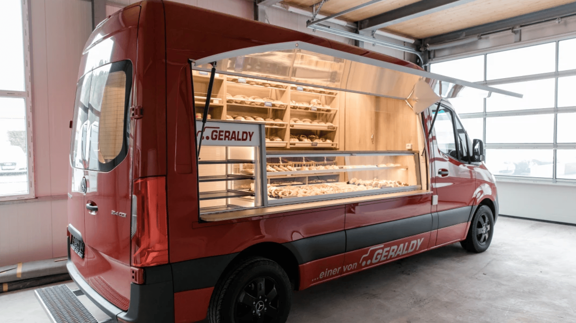 A red food truck with a large side window displays shelves filled with various baked goods, such as bread and pastries, parked inside a garage with natural light coming through the windows.