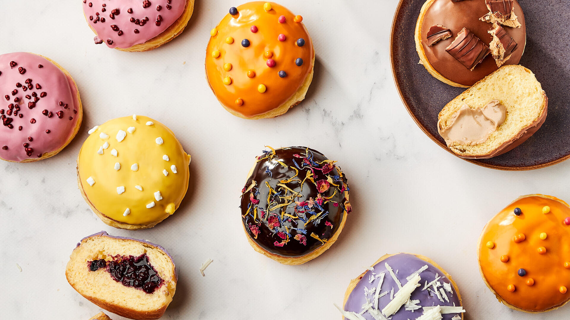 Colorful, glazed doughnuts with different fillings and toppings are arranged on a white surface, with one doughnut cut open on a plate to reveal a creamy filling.