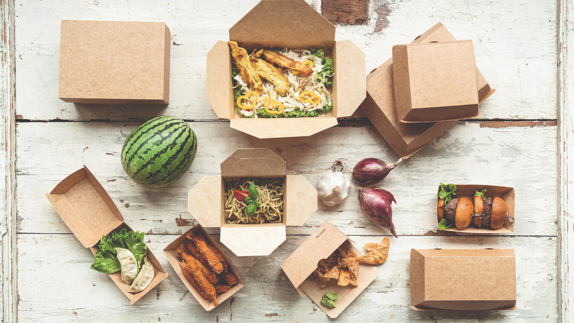 Gemischtes Essen zum Mitnehmen in umweltfreundlichen Pappbehältern auf einem rustikalen Holztisch, mit frischen Zutaten wie einer Wassermelone, Knoblauch und Zwiebeln.