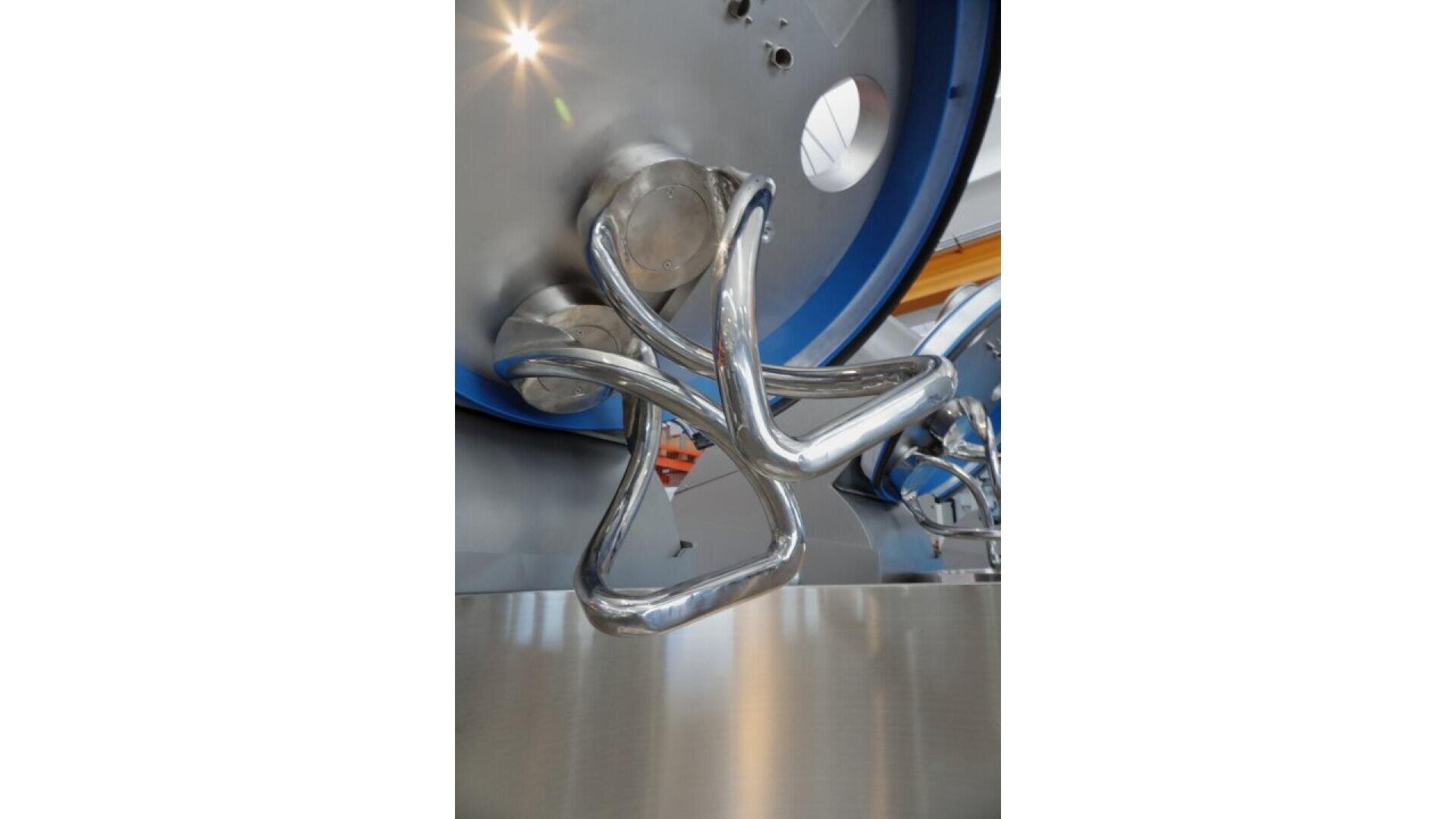 Nahaufnahme einer glänzenden, metallischen industriellen Mischschaufel mit verdrehten Armen, die an einer großen runden Maschine befestigt ist, die wahrscheinlich für die Lebensmittelverarbeitung oder -herstellung verwendet wird. Der Hintergrund ist unscharf mit Andeutungen von Maschinen.