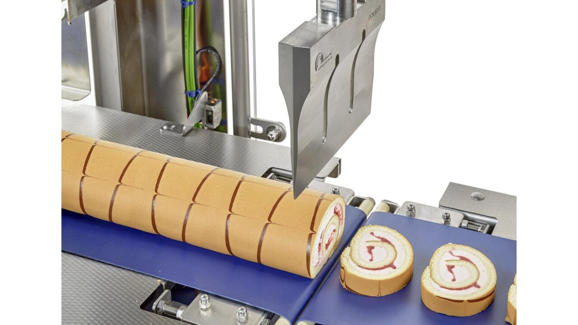 A machine with a large metal blade slices a rolled sponge cake with cream and fruit filling into uniform pieces on a conveyor belt.