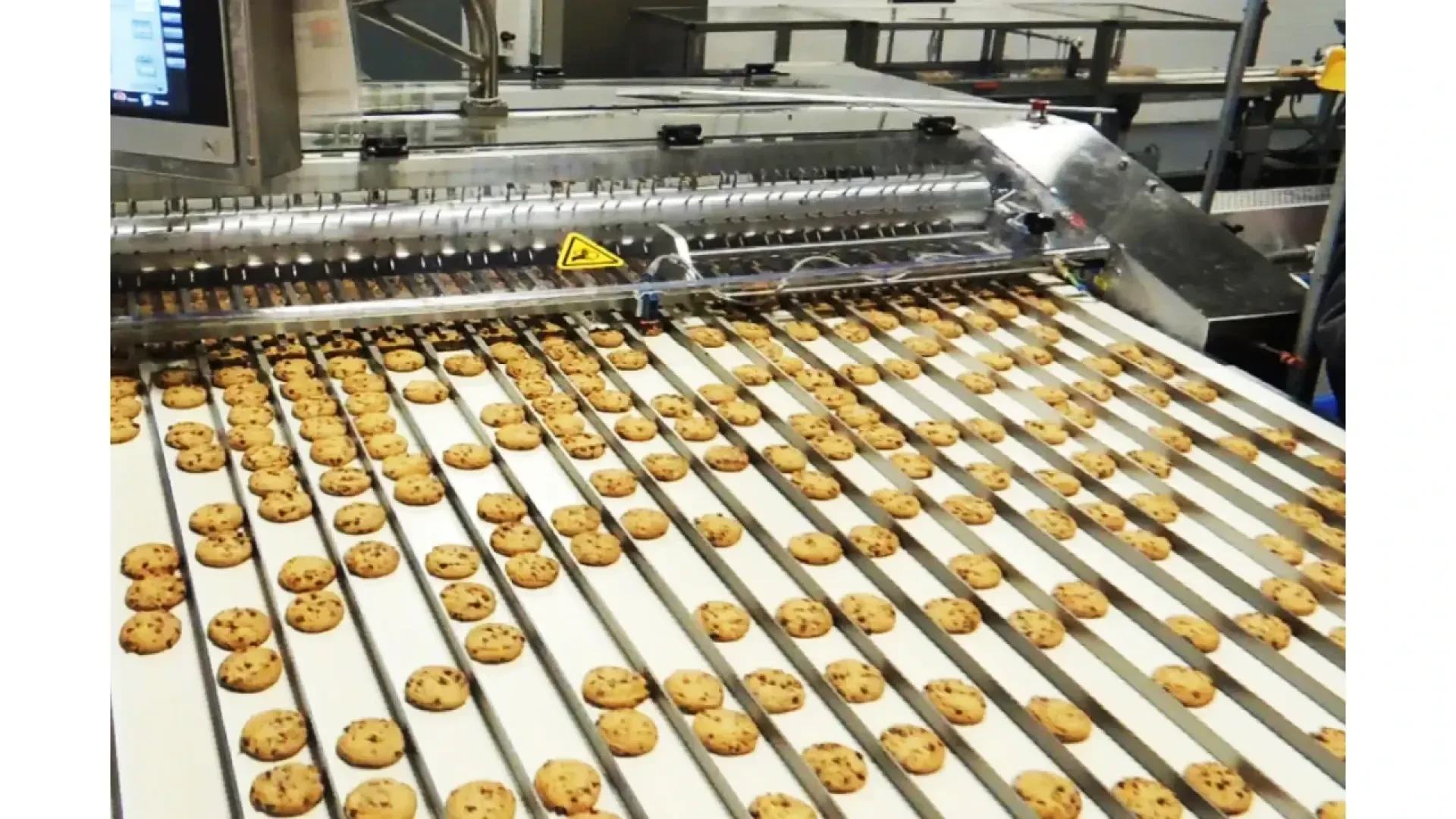 Rows of chocolate chip cookies move along conveyor belts in a factory, passing through a large stainless steel machine for processing or packaging.