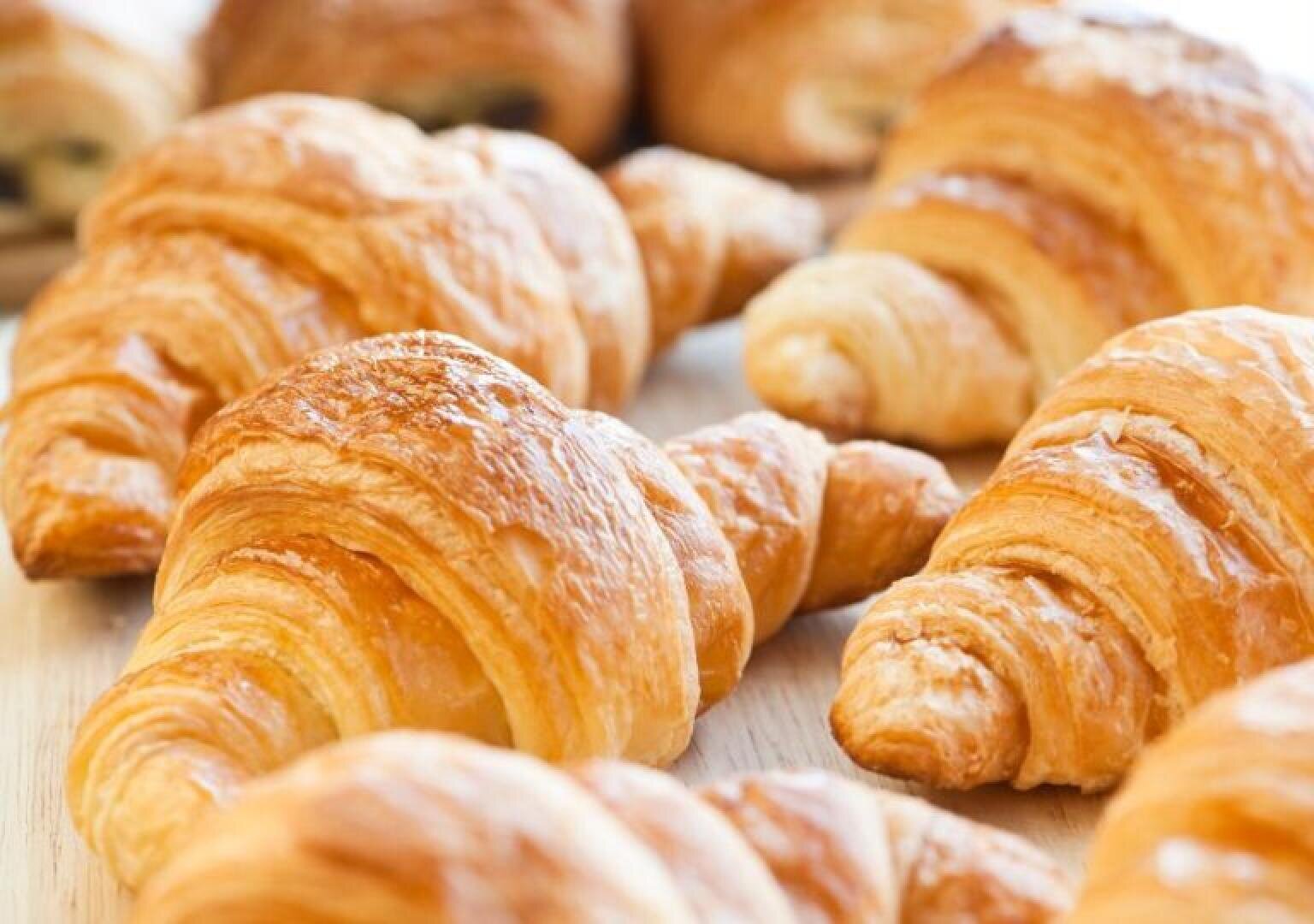 A close-up of several golden, flaky croissants arranged on a light wooden surface, showcasing their crisp, layered texture.