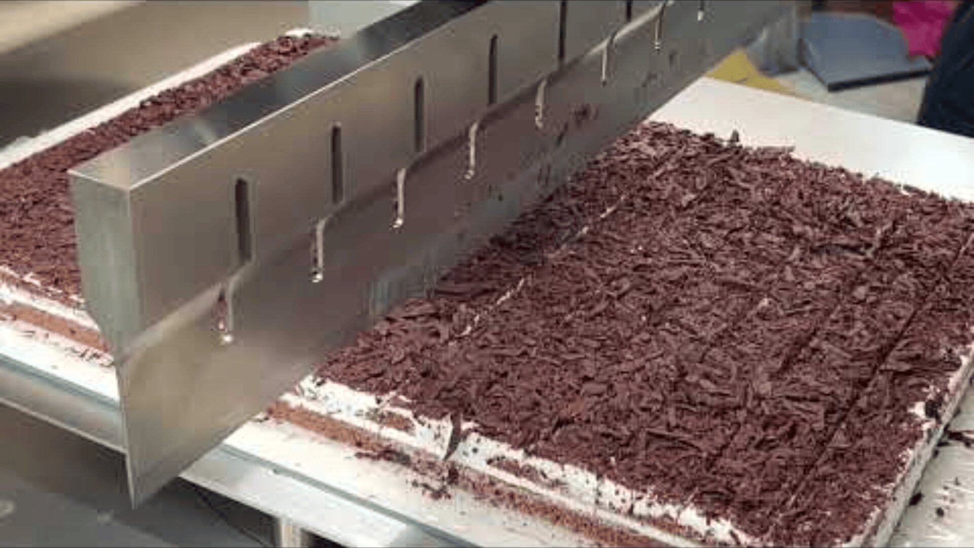 Ein großes Metallmesser schneidet einen rechteckigen Kuchen mit Schokoladenraspeln in gleichmäßige Stücke auf einer Metallfläche.