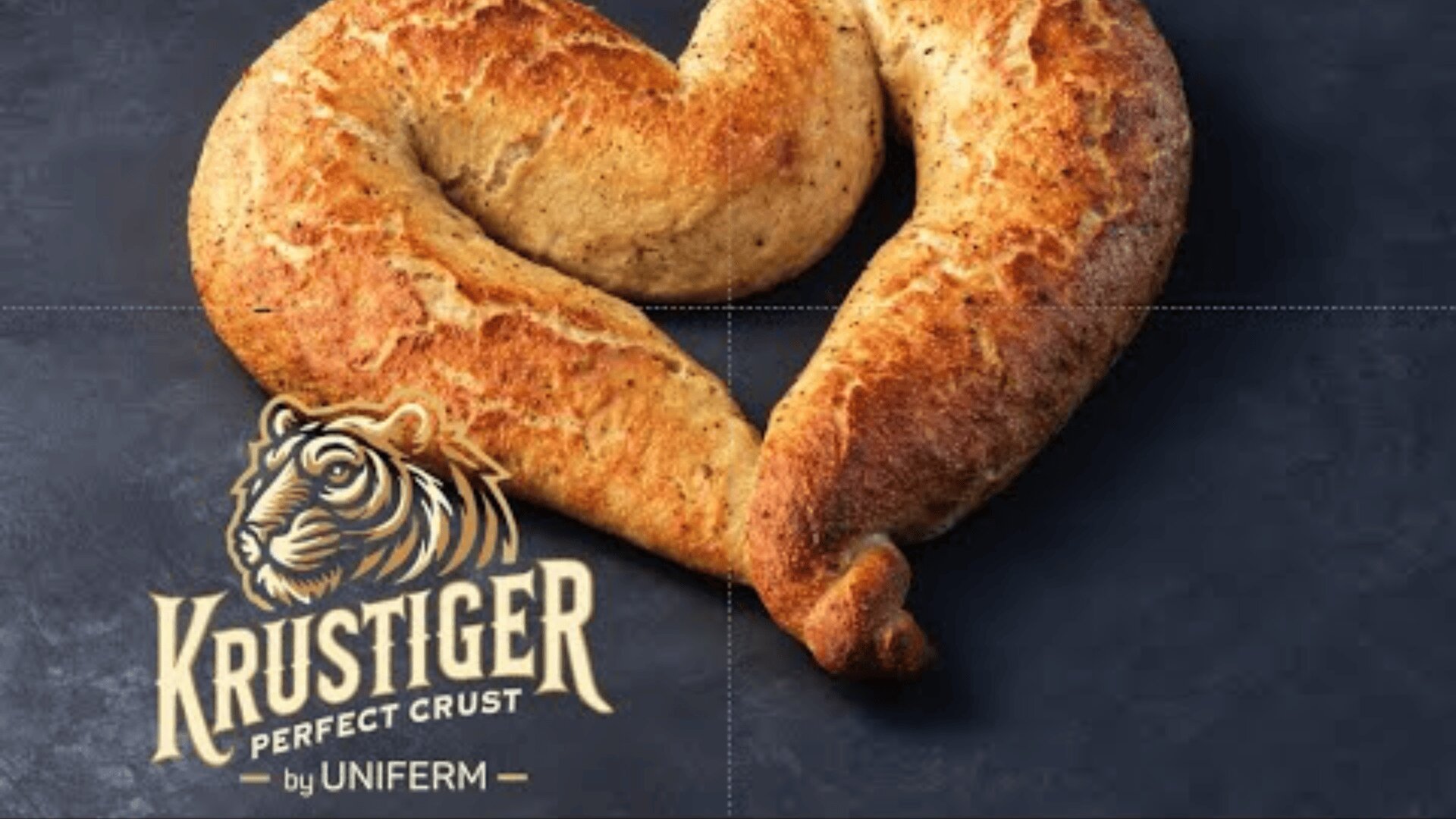 Ein herzförmiger Laib knuspriges Brot auf einer dunklen Oberfläche, mit dem Logo „Krustiger Perfect Crust“ von Uniferm, einem Tiger in der unteren linken Ecke.