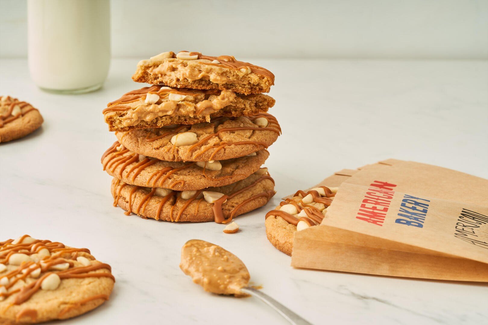 A stack of peanut butter cookies with white chocolate chips and a drizzle of peanut butter, next to a spoonful of peanut butter, a paper bag labeled American Bakery, and a glass of milk in the background.