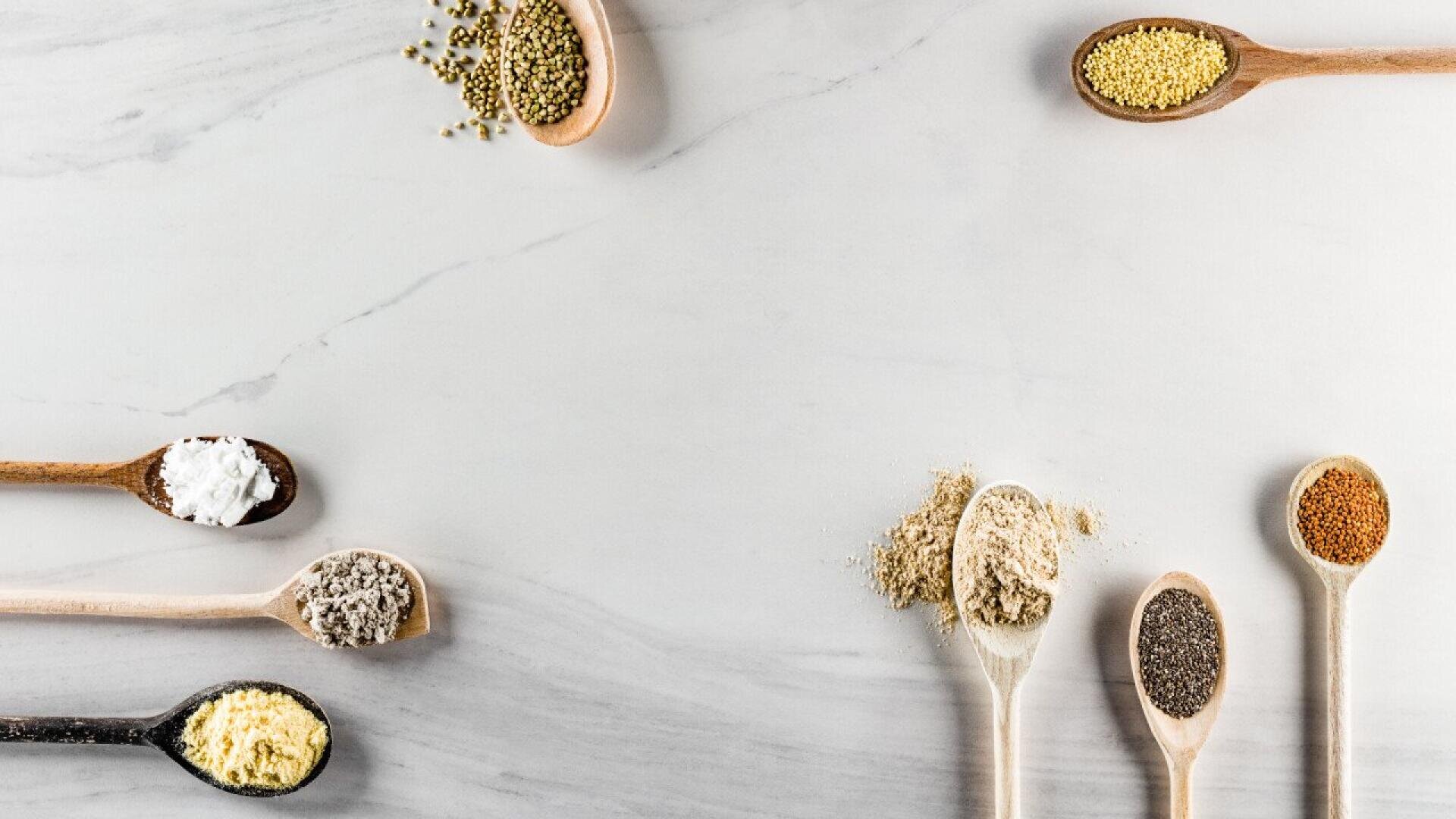 Various wooden spoons with assorted spices and powders are arranged along the edges of a light marble surface, leaving the center empty for copy space. The ingredients include seeds, granules, and powders in different colors.