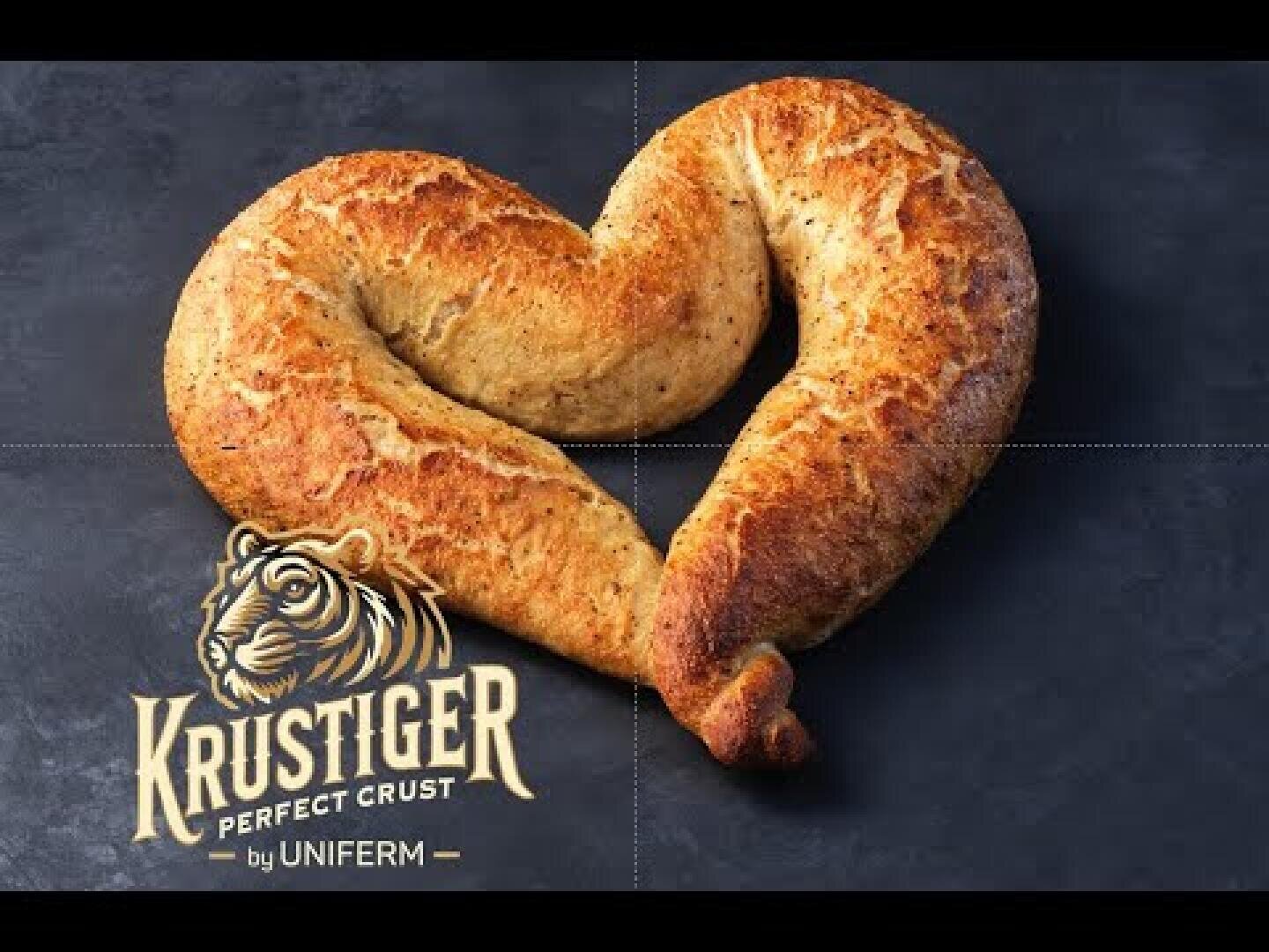 A loaf of bread shaped like a heart sits on a dark surface. The logo “KRUSTIGER PERFECT CRUST by UNIFERM” with a tiger image appears in the lower left corner.