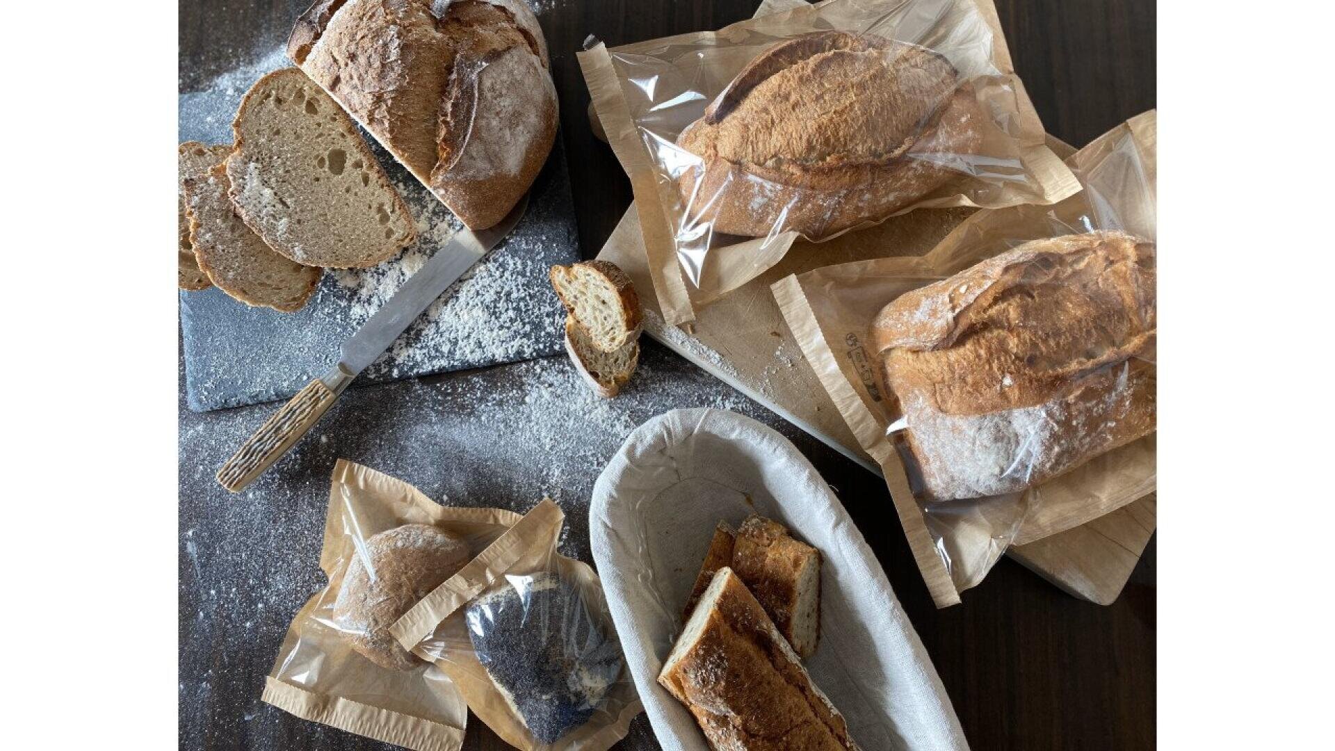 Auf einem dunklen Tisch liegen handwerklich hergestellte Brotlaibe, teilweise in Scheiben geschnitten, mit einem Brotmesser, Papiertüten, Krümeln und Mehl verstreut. Mehrere Brotstücke und ganze Brote sind teilweise in Papier eingewickelt.