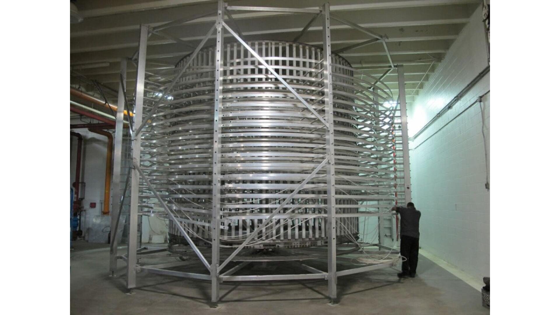Eine große, zylindrische Metallstruktur mit spiralförmigen Regalen ist in einem Industrieraum untergebracht. Eine Person steht in der Nähe, um die beeindruckende Höhe und den komplexen, vielschichtigen Aufbau der Maschine zu verdeutlichen.