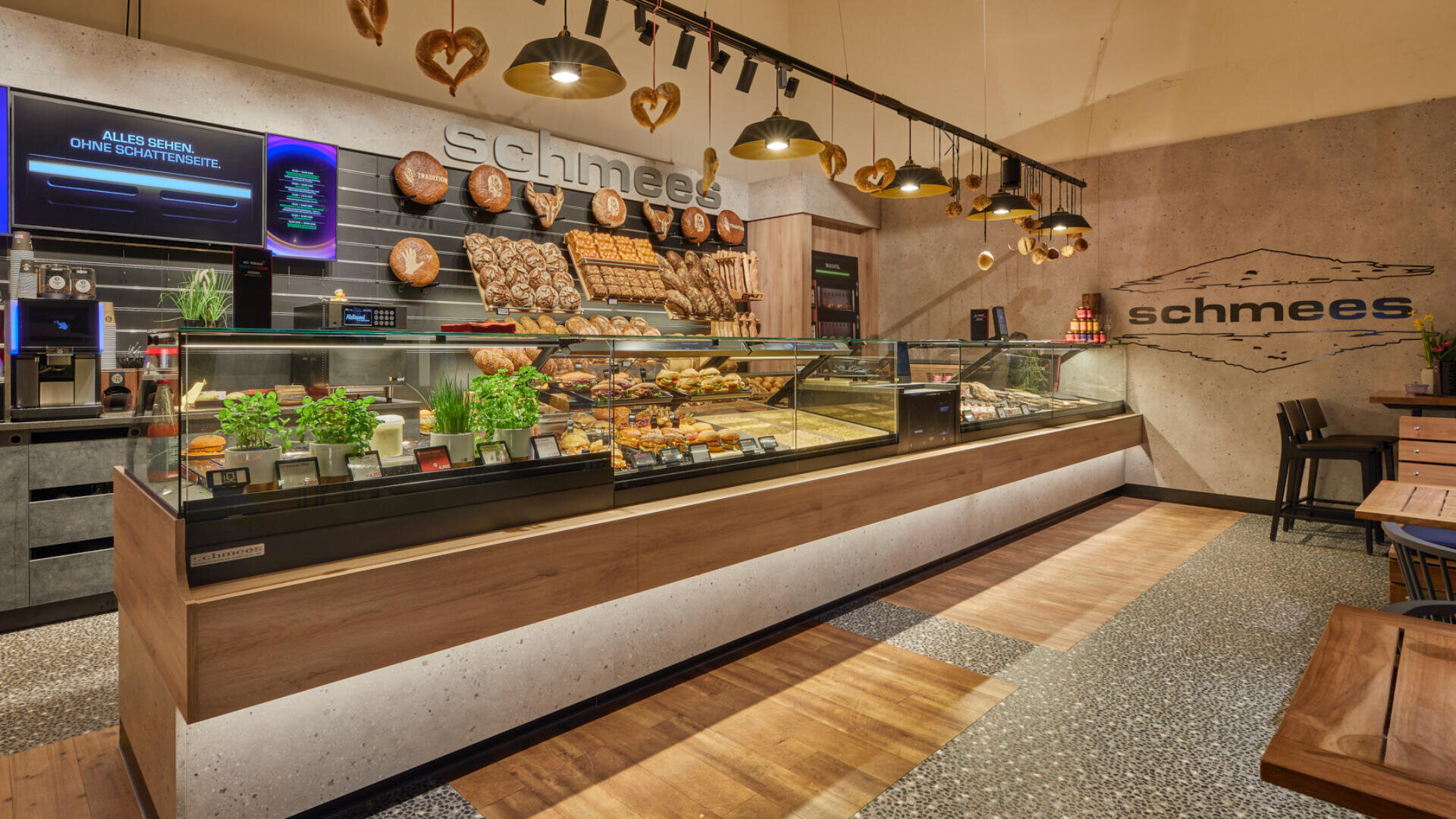 Moderne Bäckerei mit Holzakzenten, Glasvitrinen mit Brot, Gebäck und Kräutern. Hängelampen und Brezeln schmücken den Raum. An der Wand steht der Name schmees.