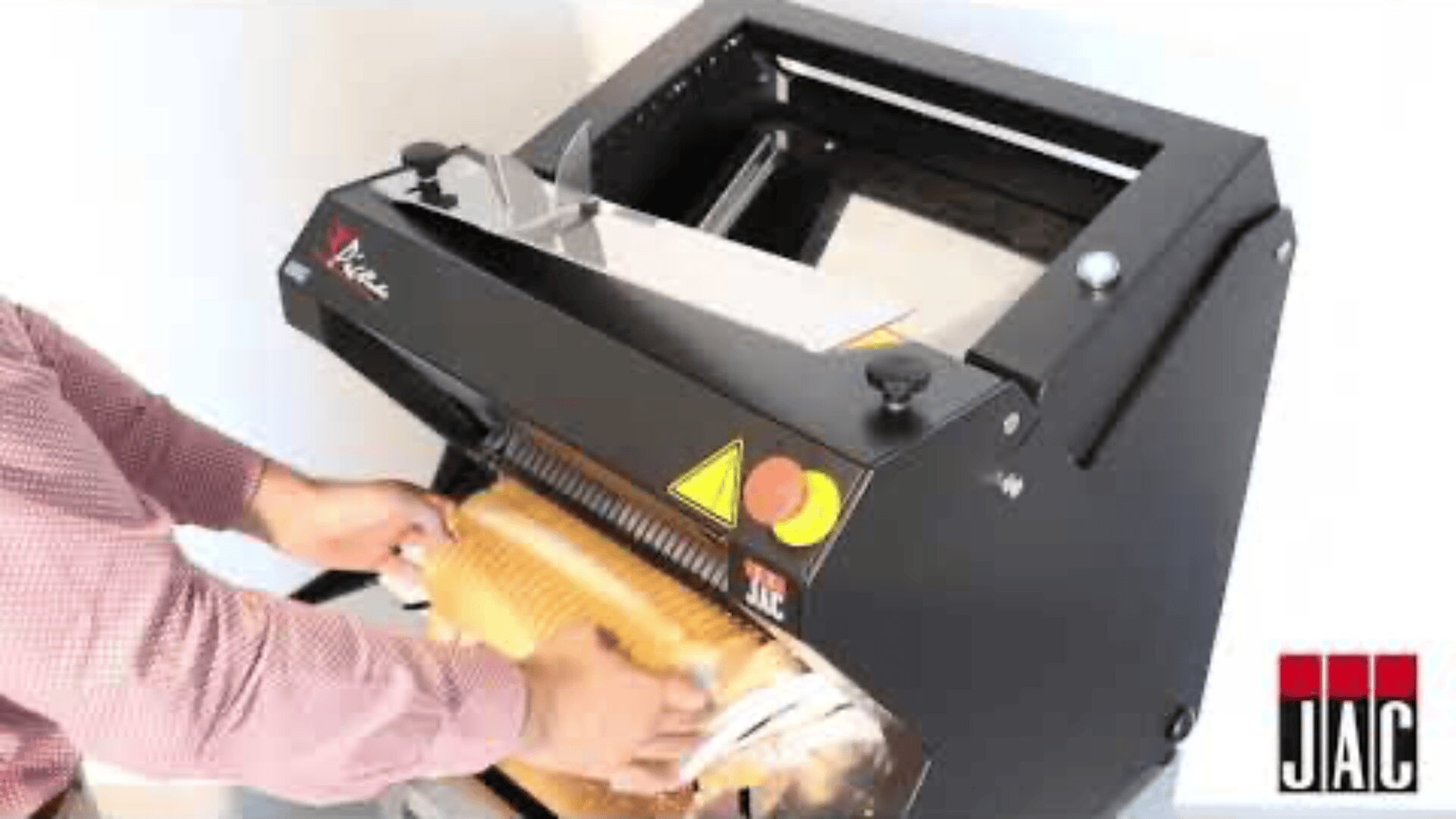 A person is placing a loaf of bread into a commercial bread slicing machine. The machine is black with visible control buttons and the JAC logo in the bottom right corner.