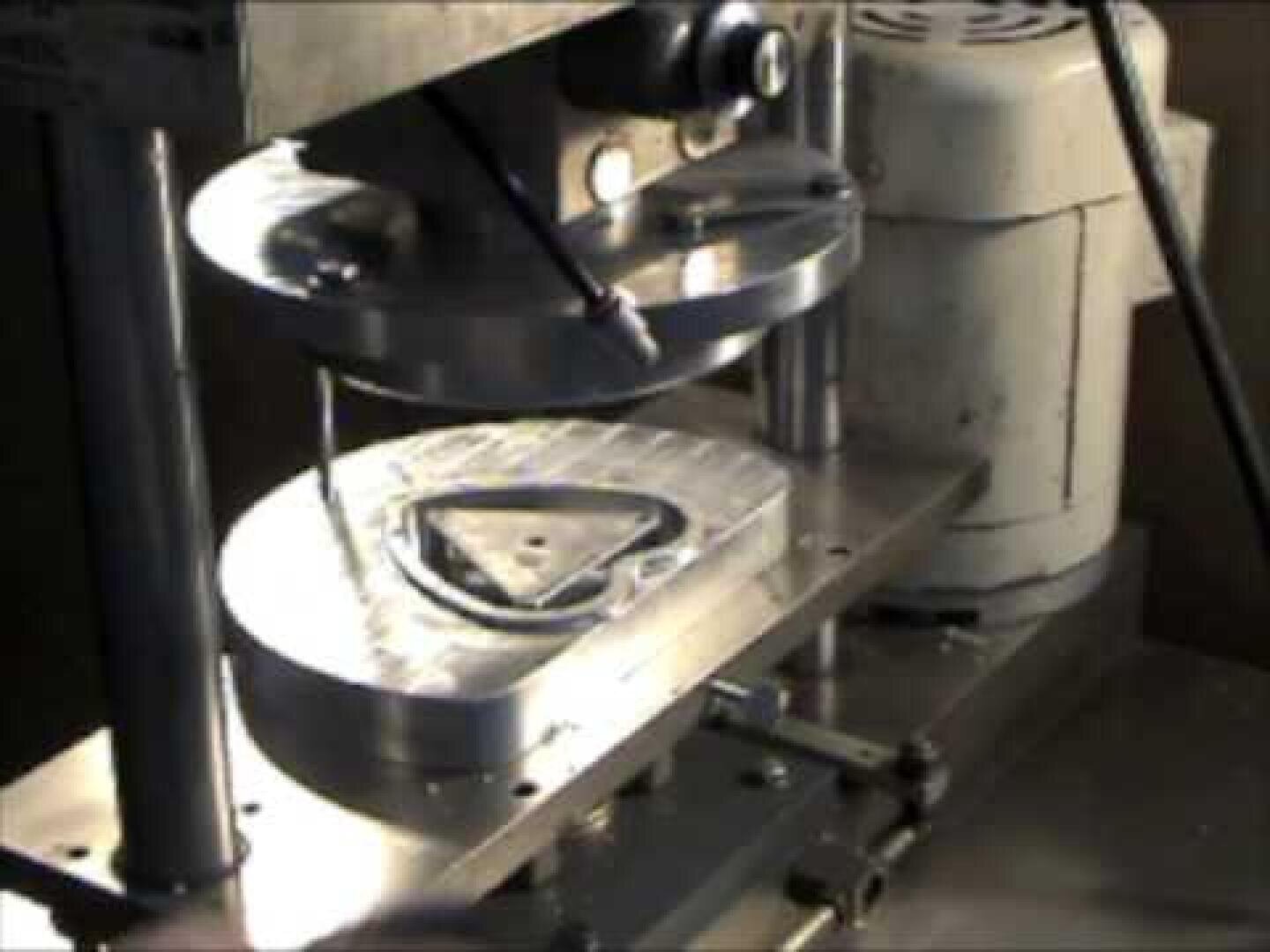 A close-up of a metal forming machine pressing a triangular piece of metal between two circular dies, with mechanical components and a motor visible in the background.