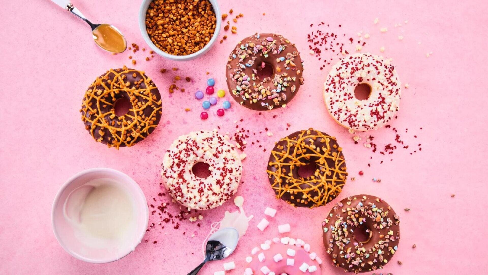 Six decorated donuts with colorful sprinkles and toppings are arranged on a pink surface, surrounded by bowls of icing, a spoon with caramel, scattered candies, and mini marshmallows.
