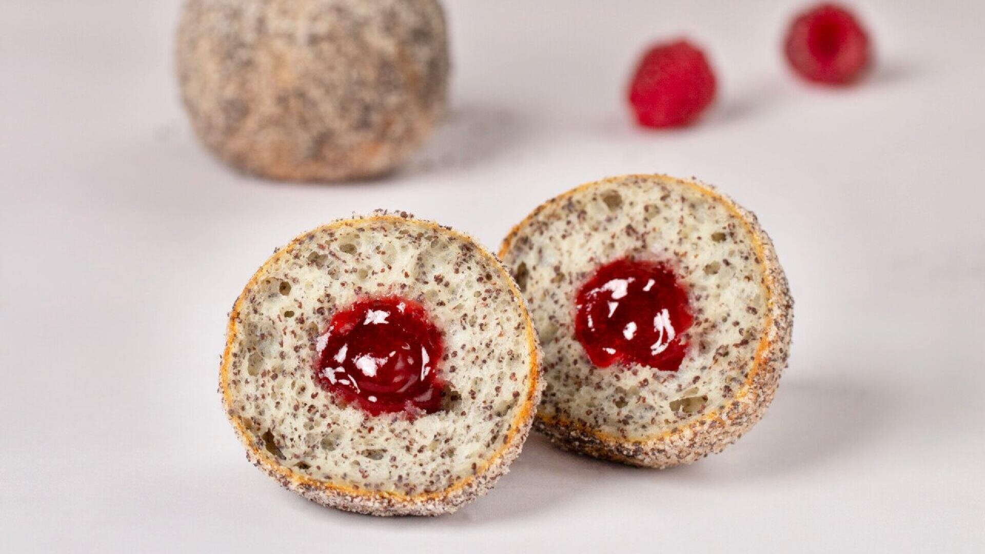 A poppy seed pastry cut in half, revealing a bright red raspberry jam filling, with another whole pastry and two raspberries in the blurry background on a light surface.