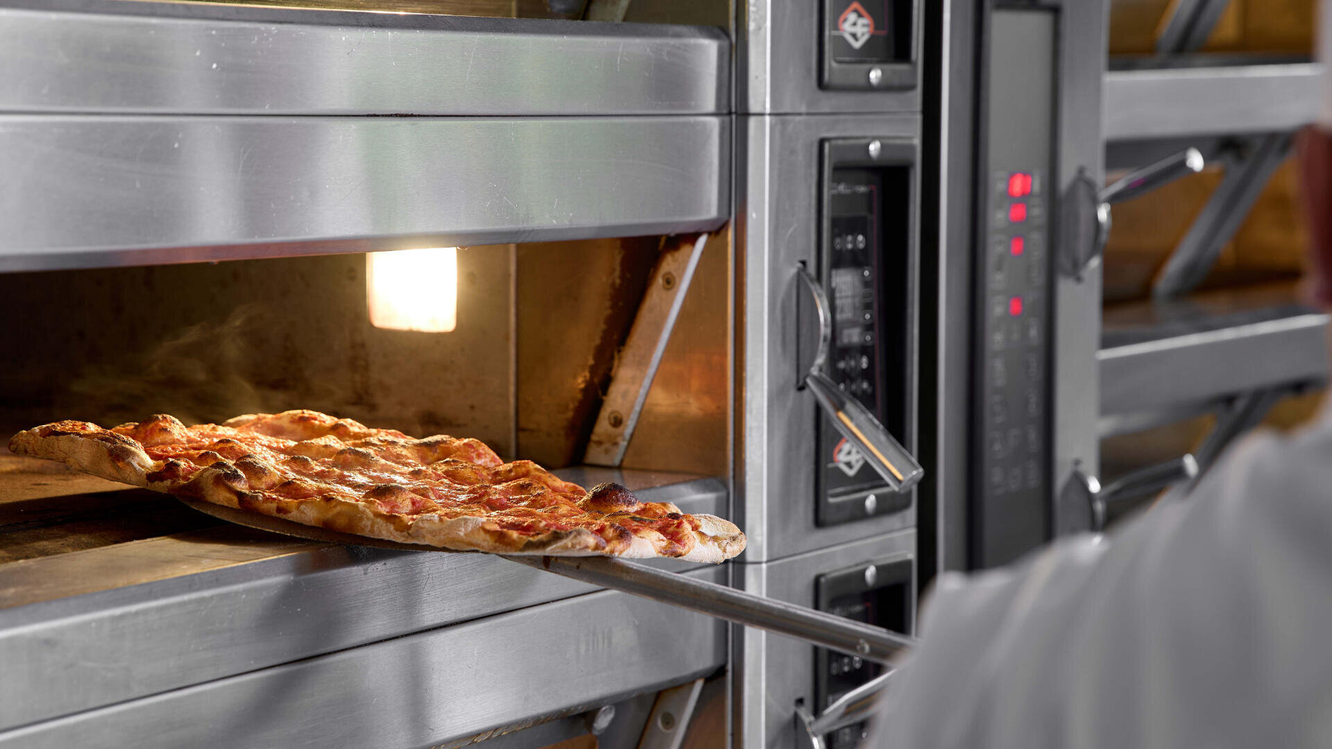 A person uses a pizza peel to place or remove a freshly baked pizza from a stainless steel commercial oven, with the oven interior warmly lit.