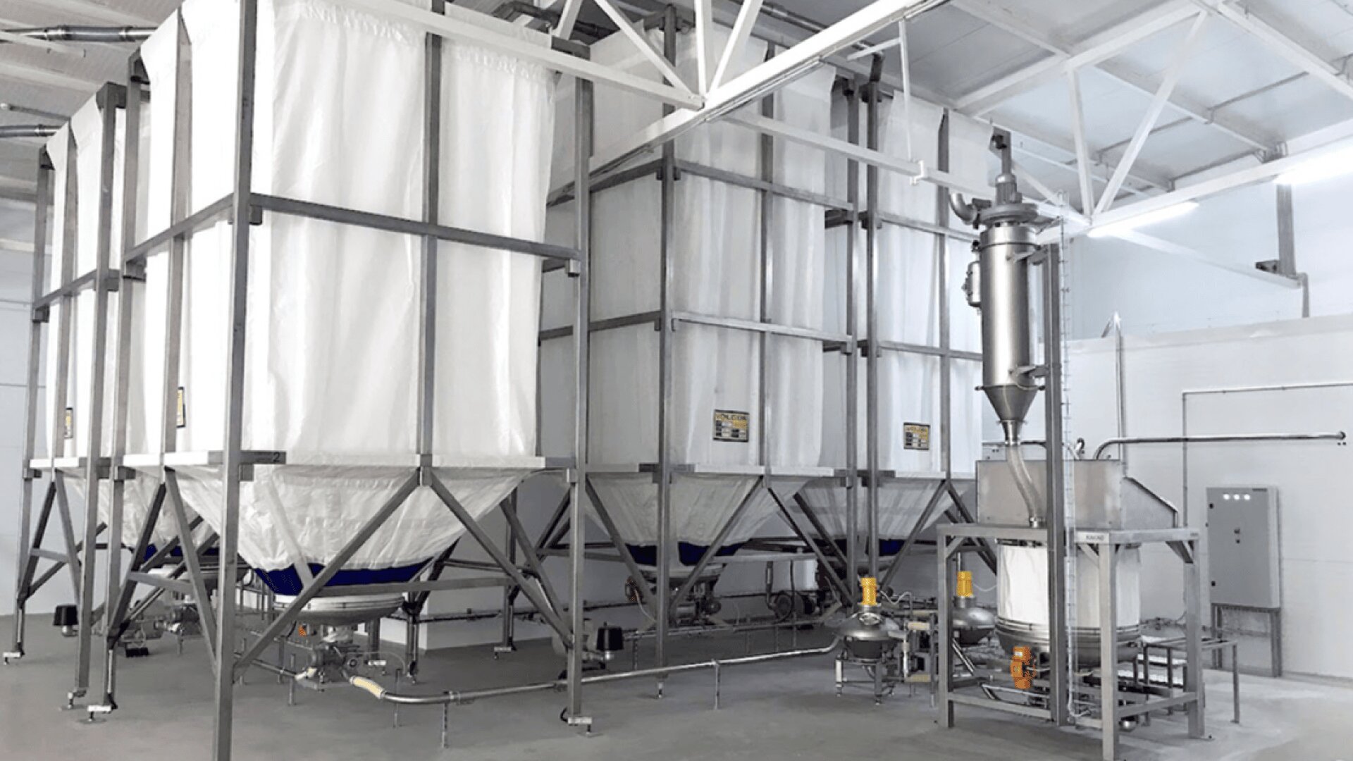 Große Industriesilos mit weißen Stoffabdeckungen werden von Metallrahmen in einer sauberen, modernen Anlage getragen. Rohre, Maschinen und Bedienfelder sind entlang des Bodens und der Wände sichtbar.