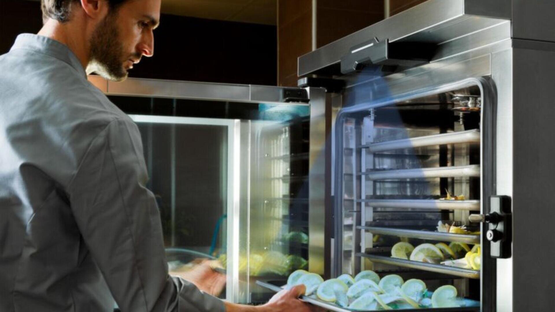 A chef places trays of food into a commercial stainless steel oven in a professional kitchen. The oven has multiple shelves with evenly spaced trays containing food items.