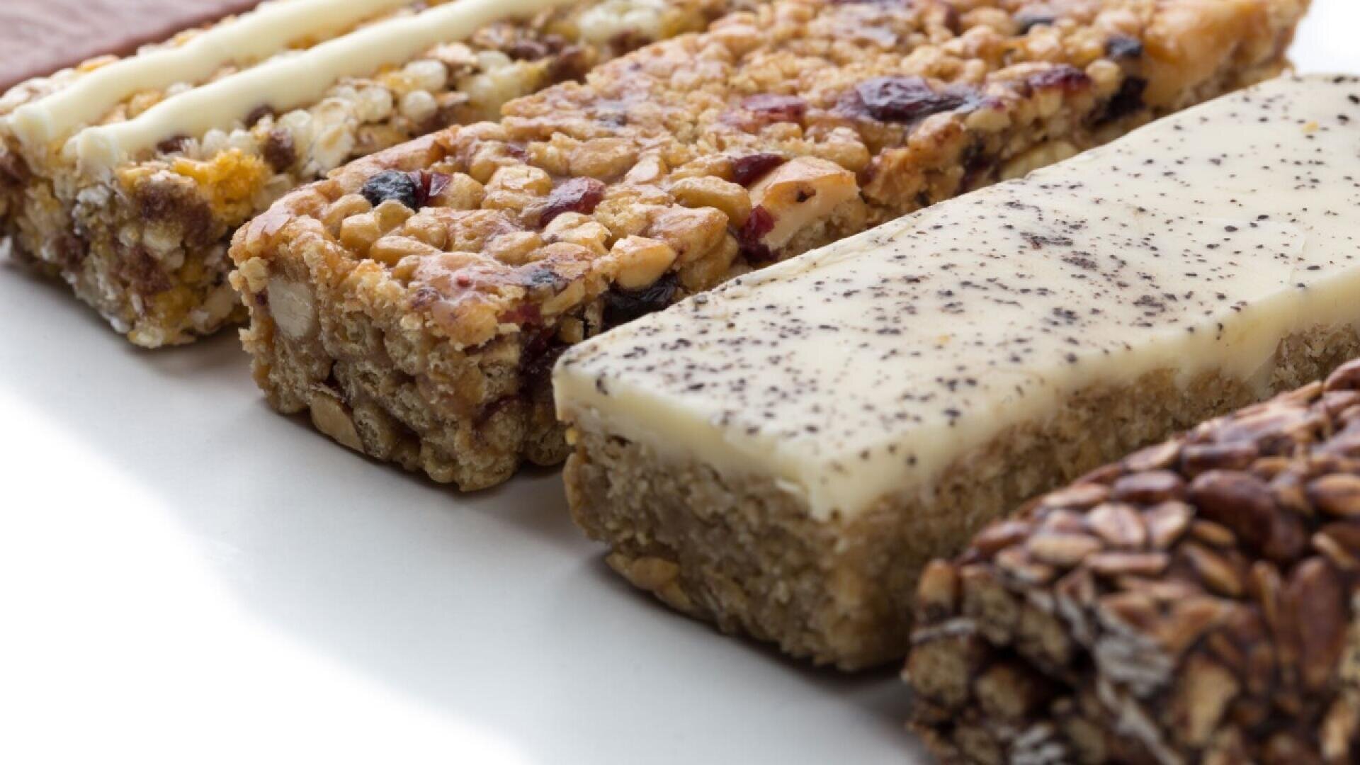 Four assorted granola bars with various toppings, including nuts, dried fruits, and different types of coatings, arranged side by side on a white surface.
