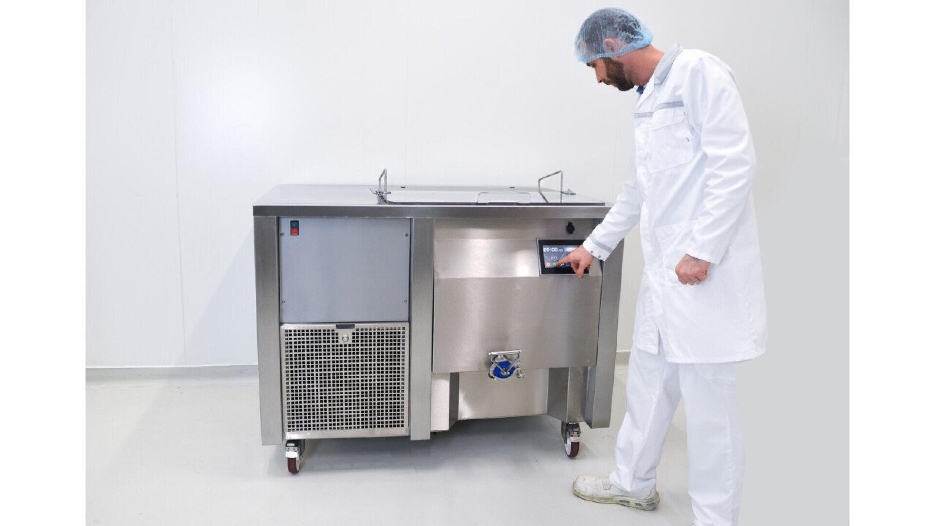 Eine Person in weißem Laborkittel, Haarnetz und Schuhüberziehern bedient in einer sauberen, sterilen Umgebung eine große Industriemaschine aus rostfreiem Stahl mit einem Bedienfeld.