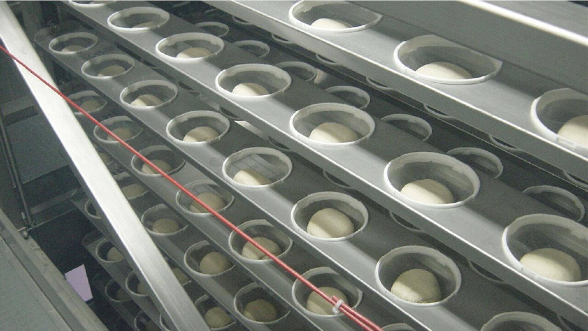 Reihen von Teigkugeln, die in einzelnen runden Fächern auf einem mehrstöckigen Metallgestell angeordnet sind, möglicherweise in einer industriellen Brot- oder Bäckereiproduktionsanlage.