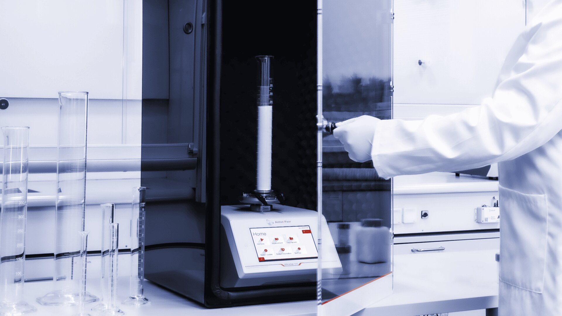 A scientist in a white lab coat operates laboratory equipment, placing a graduated cylinder with white powder inside a machine. More graduated cylinders are on the counter, and the lab has a clean, modern setup.