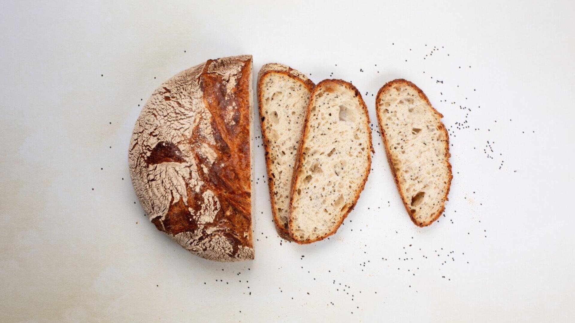 A round loaf of rustic bread, partially sliced, with three slices arranged next to it on a light surface. A few scattered poppy seeds are visible nearby.