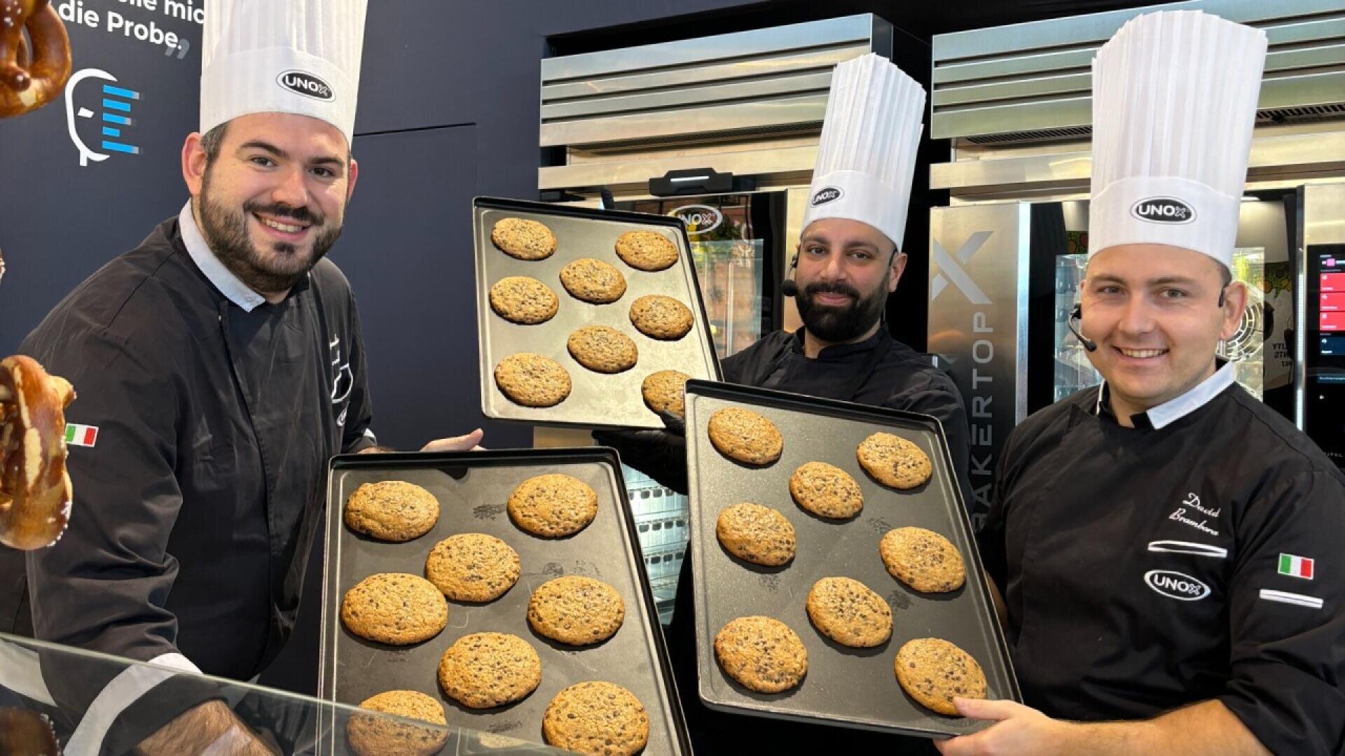 Three smiling bakers in tall white hats and black uniforms hold trays of large chocolate chip cookies, standing in a modern bakery kitchen.