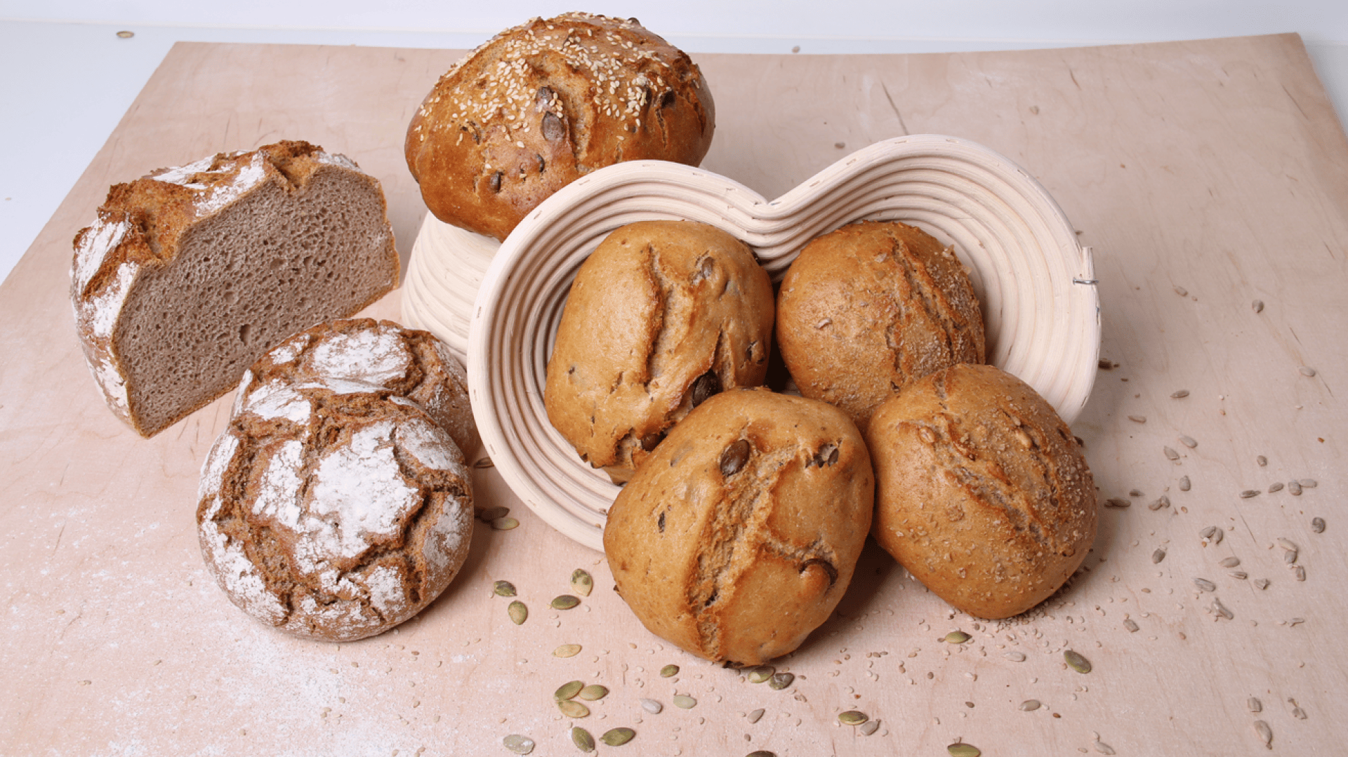 A variety of rustic breads, including round rolls and a sliced loaf, are displayed on a wooden surface with some pumpkin seeds scattered around and a beige bread basket holding several rolls.