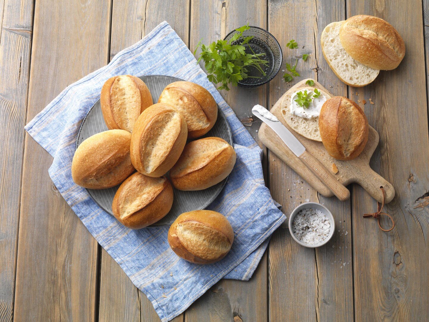 Ein Teller mit runden Brötchen auf einem blauen Tuch über einem Holztisch, mit einem aufgeschnittenen Brötchen auf einem Schneidebrett, einem Messer, einer kleinen Schüssel mit Salz, Kräutern und einer kleinen Schale mit Frischkäse in der Nähe.